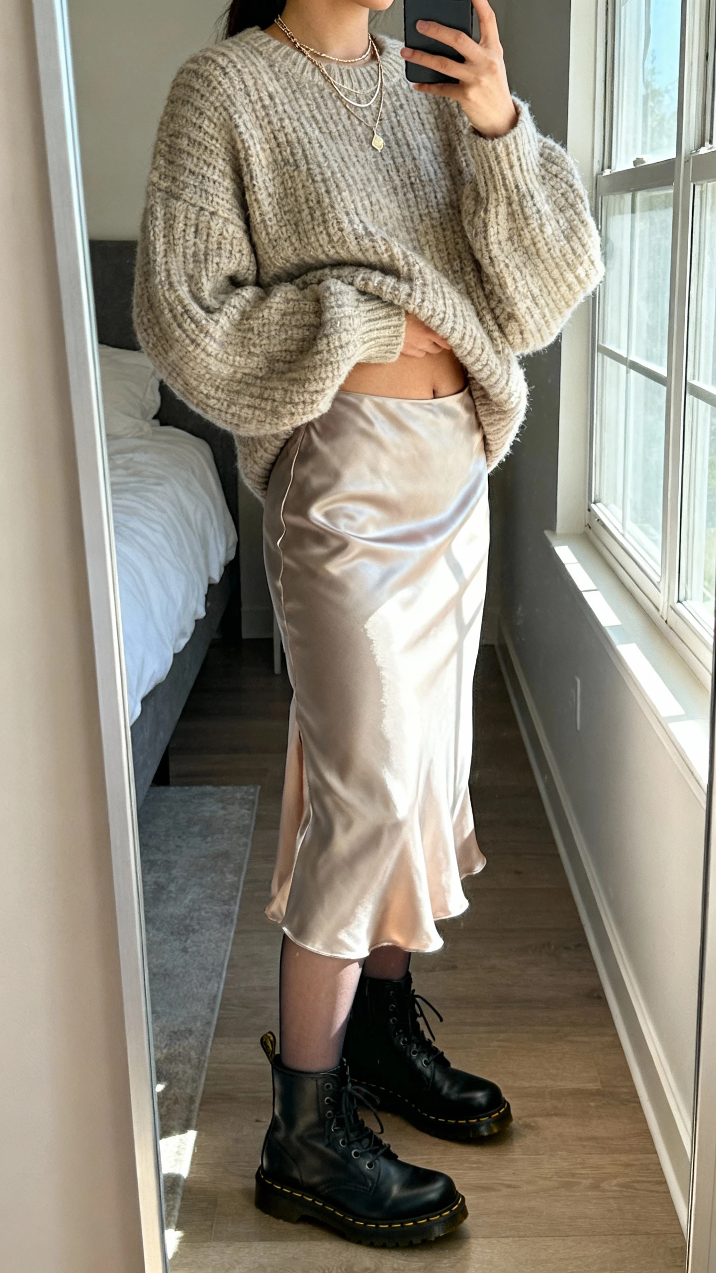 iPhone side-angle mirror selfie of a woman wearing a satin slip skirt, chunky oversized knit sweater half-tucked, semi-opaque tights, black Doc Martens boots, and delicate layered necklaces, face not visible, bedroom mirror with natural window light, iPhone photo quality.