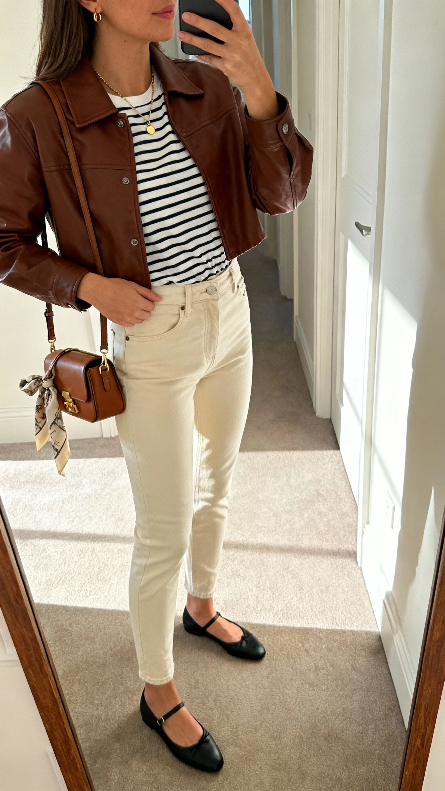 Side-angle iPhone mirror selfie of a woman wearing a cropped chocolate-brown leather jacket, Breton striped tee, high-waisted ecru slim jeans, black ballet flats, mini satchel, delicate gold necklace and tiny hoops, optional silk scarf tied to bag; face not visible, cropped at shoulders, hallway full-length mirror with natural light.