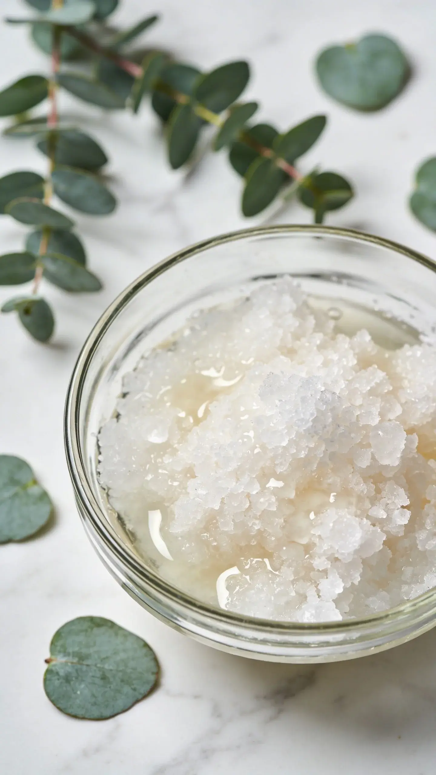 Overhead close-up shot of coarse white sea salt crystals mixed with coconut oil in a glass bowl, with fresh eucalyptus sprigs and scattered eucalyptus leaves on a white marble surface, natural lighting, spa aesthetic, shallow depth of field focusing on the crystalline texture of the salt scrub
