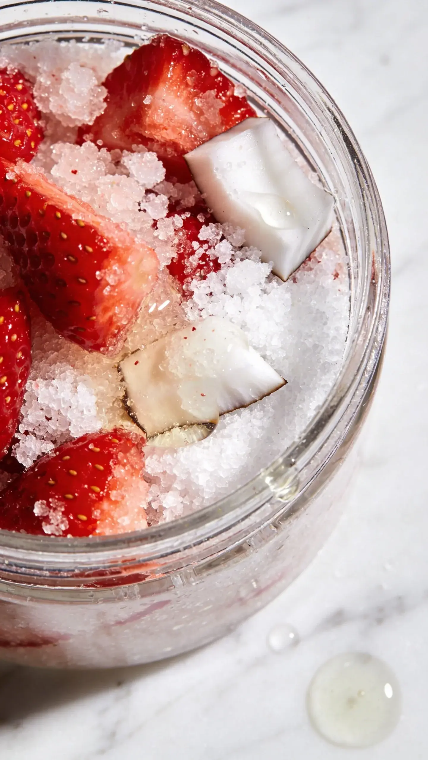 Extreme close-up of fresh red strawberries mixed with white granulated sugar and coconut oil in a clear glass jar, showing the texture of the scrub with visible sugar crystals, strawberry pieces, and oil droplets, shot from above with natural lighting on a white marble surface