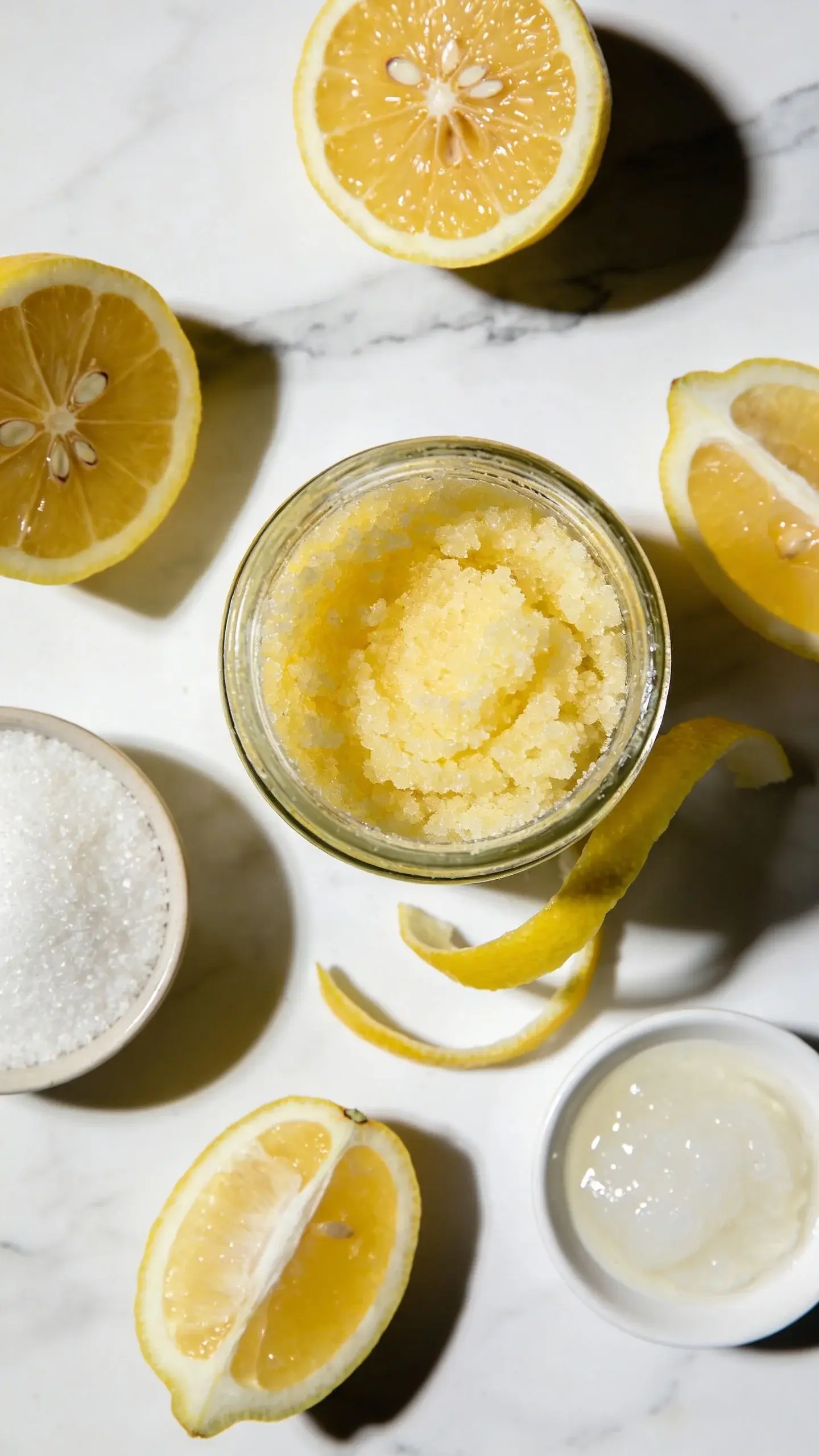 Overhead close-up photo of a glass jar filled with yellow-tinted sugar scrub, surrounded by fresh lemon halves, granulated white sugar in a small bowl, and coconut oil, arranged on a white marble surface with natural lighting casting soft shadows