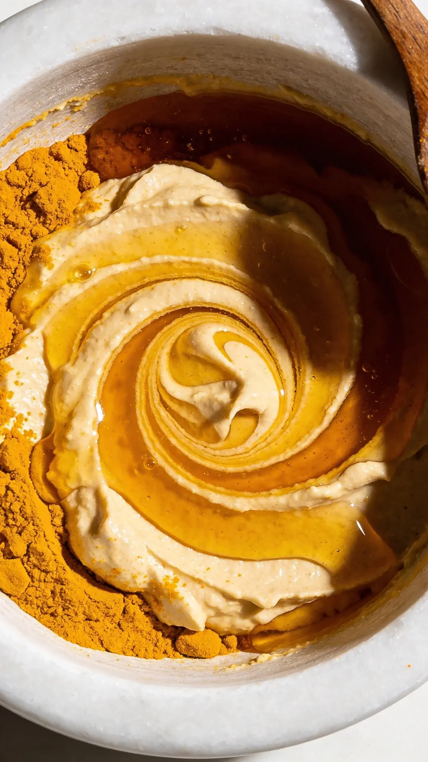 Extreme closeup overhead shot of golden turmeric powder and thick amber honey swirled together in a white ceramic bowl, creating marbled patterns with rich golden and deep yellow tones, natural lighting casting soft shadows, showing the creamy texture of the freshly mixed bath scrub with a wooden spoon partially visible at the edge