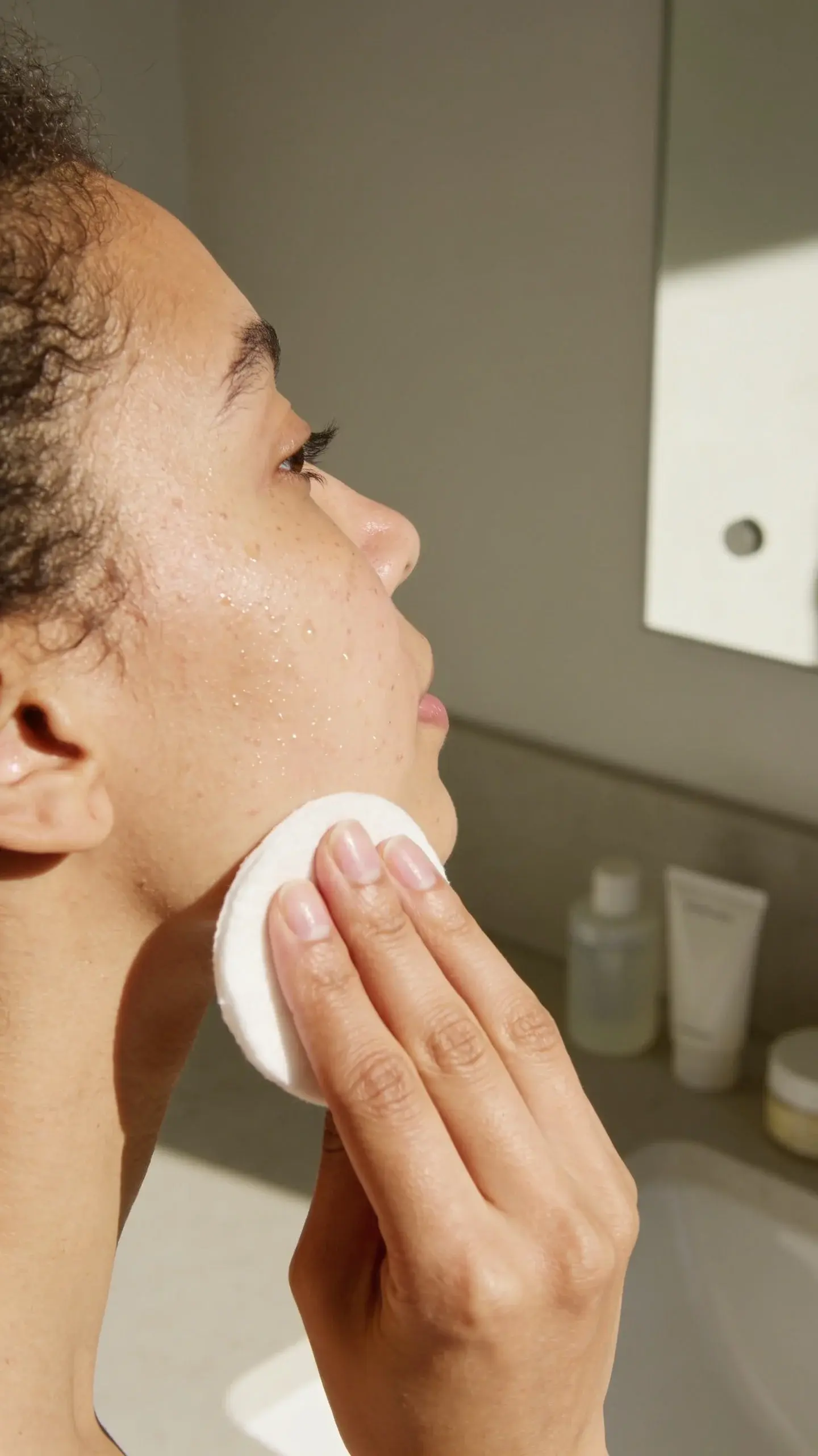 Serene bathroom vanity scene capturing the moment of hydration: model with naturally dry, slightly flaky skin pressing a cotton pad soaked in toner onto her cheek, visible glow and micro-droplets on skin; in the foreground, an open bottle of alcohol-free toner with a watery-gel texture next to a small bowl of water and a ceramic tray holding a dropper of glycerin-like serum; eucalyptus in a glass, folded white towel, and a blurred rain-speckled window in the background; soft, cool-toned lighting emphasizing plumpness and calm, clean aesthetic, no text.