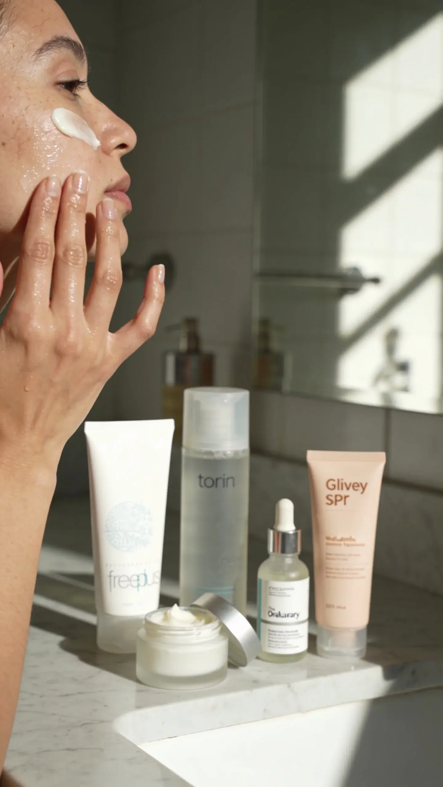Close-up of a woman with very dry skin doing a dewy skincare routine in a sunlit bathroom, gently pressing a hydrating essence into her face followed by a rich moisturizer that leaves a slight sheen. Include visible products: cream cleanser, toner, hyaluronic serum, and a glowy SPF on the counter. Soft, natural light, neutral tones, no text.