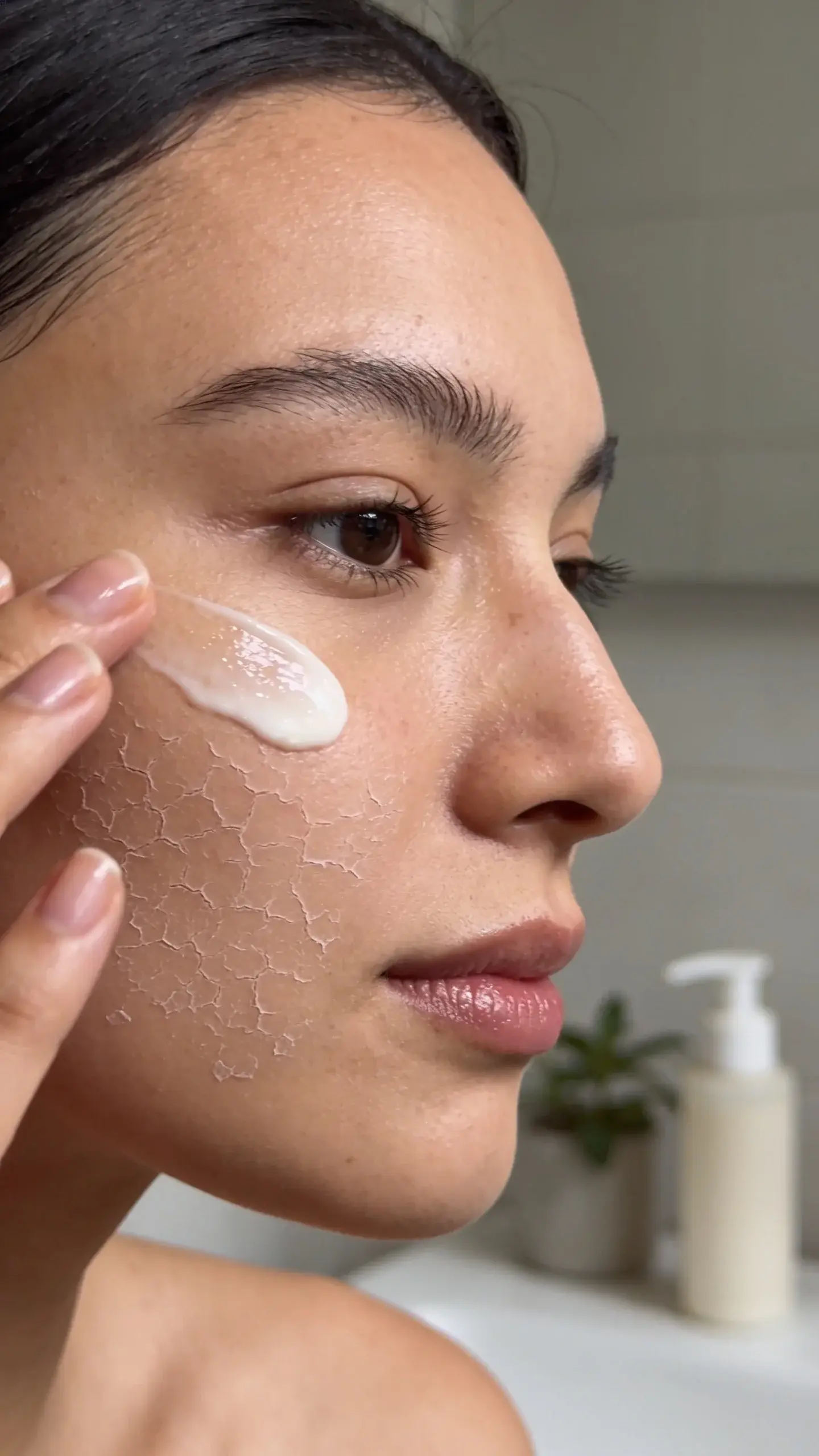 Close-up of a woman with naturally dry, flaky patches on her cheek in soft daylight, gently applying a ceramide-rich cream with visible dewy hydration on the skin. Minimal, neutral bathroom setting with a small plant and simple pump bottle in the background. Clean, realistic skincare aesthetic, no text.