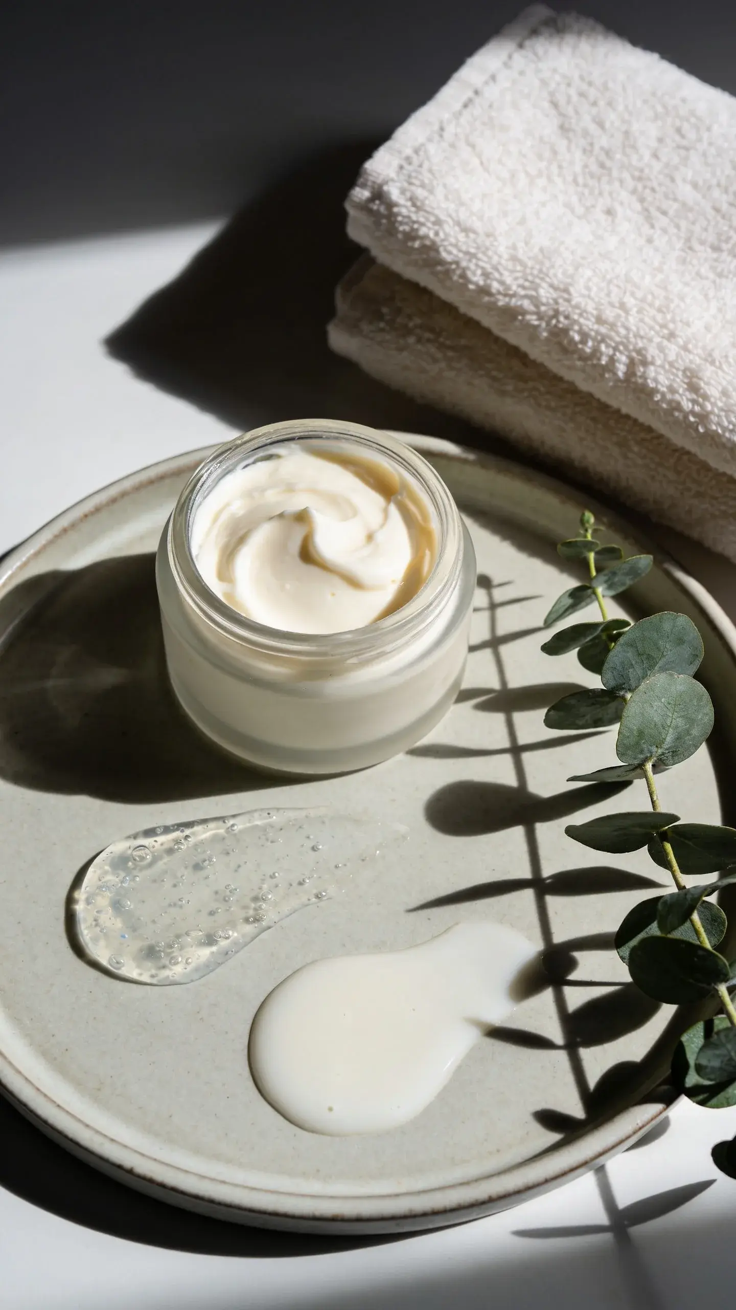 Flat lay of bathroom vanity scene featuring open jar of thick shea-butter-based cream, a translucent gel-lotion dollop showing hyaluronic texture, and a milky ceramide lotion puddle on a ceramic tray; soft morning light, eucalyptus sprig and cotton towel nearby; clean, minimal aesthetic with muted colors, no brand names or text.