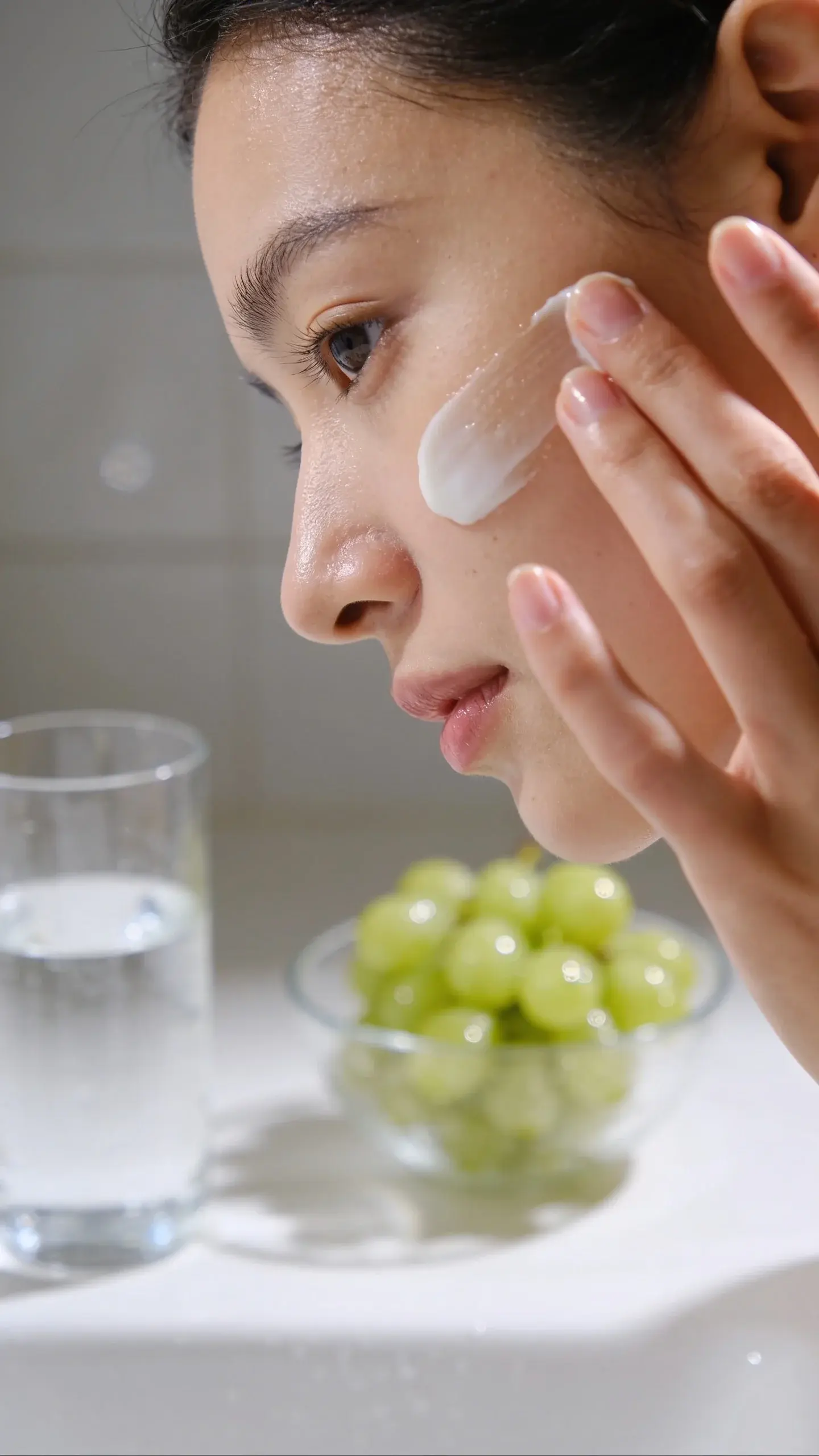 Close-up of a woman’s face with smooth, hydrated skin in soft natural light, gently applying a creamy moisturizer with fingertips; include a glass of water and a bowl of dewy grapes blurred in the background to hint at hydration. Minimal, clean bathroom setting with neutral tones and a subtle reflection.