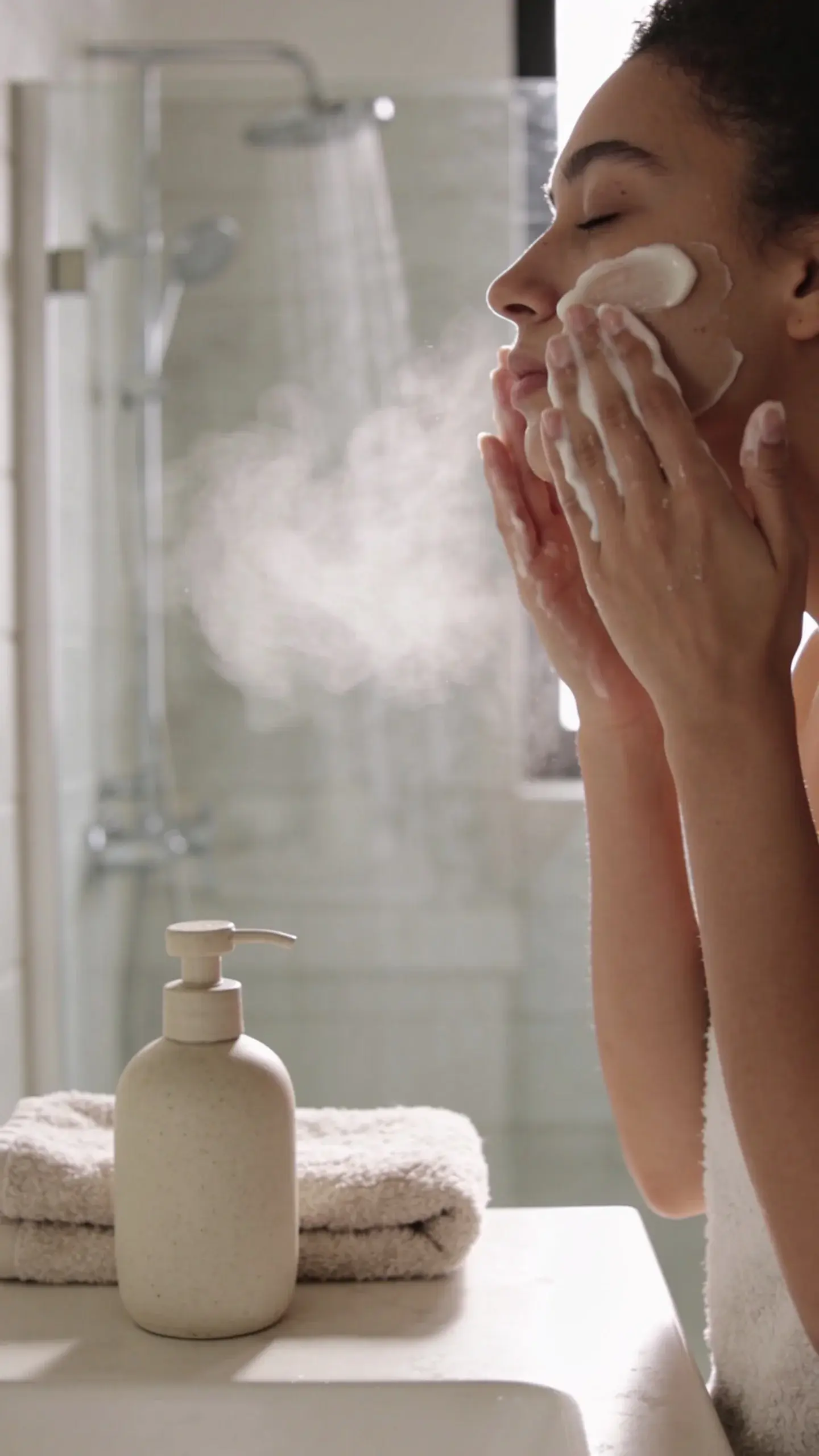 Cozy winter bathroom scene with soft morning light: a person with smooth, hydrated skin gently massaging a creamy, non-foaming cleanser onto their face, steam from a warm shower in the background, plush towel and simple ceramic pump bottle on a clean counter. Neutral tones, soft focus, serene and minimal.