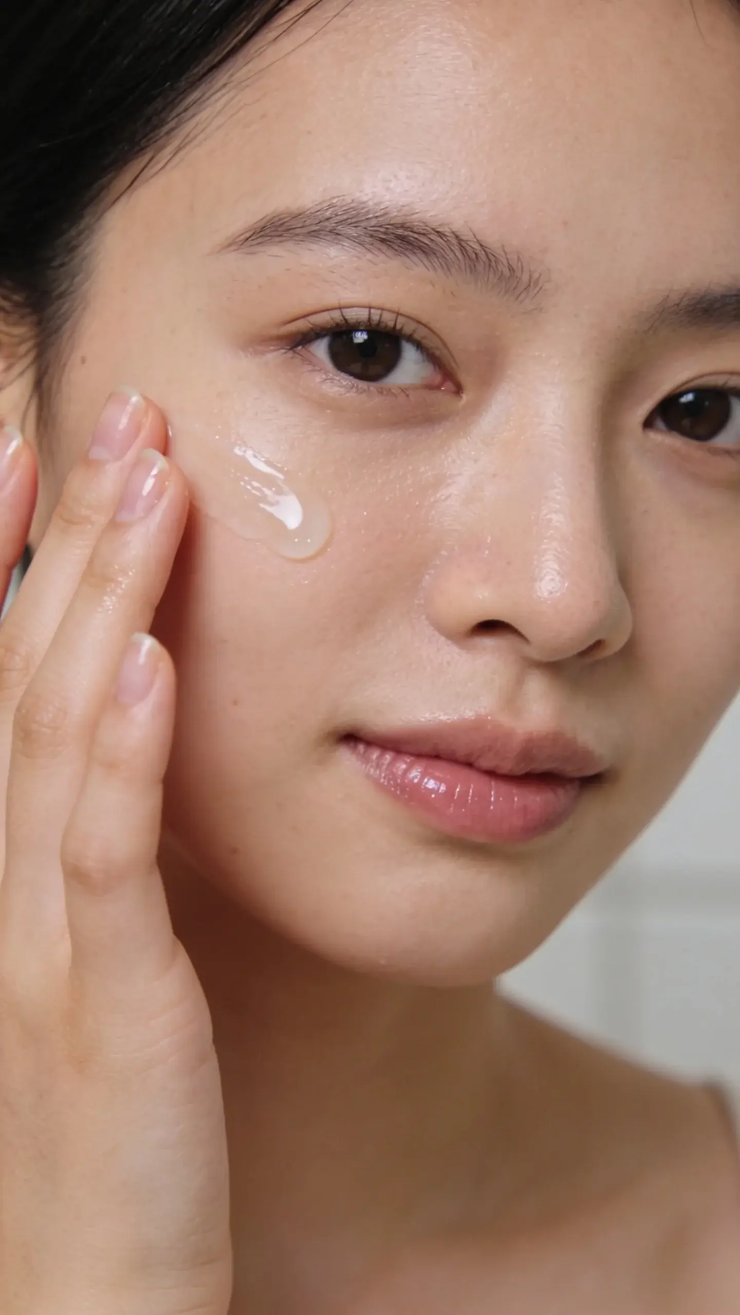 Close-up of a young person with smooth, clear, dewy skin gently pressing a lightweight gel serum into their cheek with fingertips; soft natural bathroom light, neutral background, fresh minimal vibe, no product labels visible.