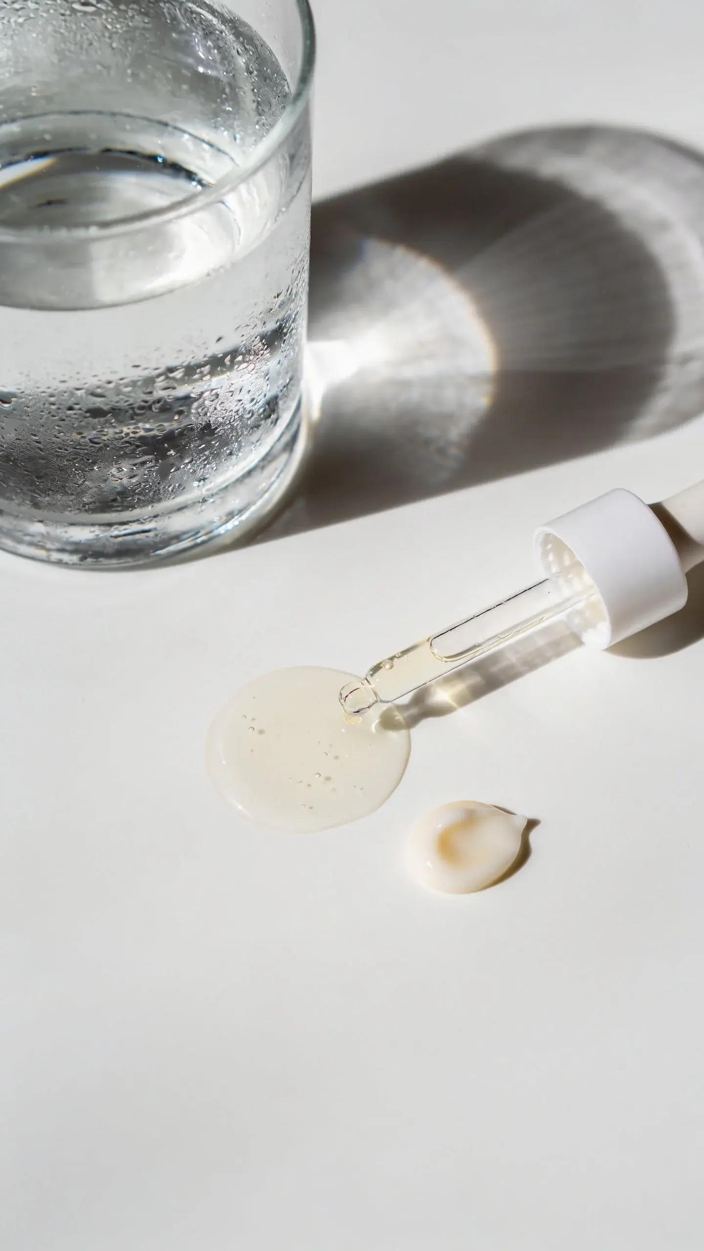 Overhead shot of a simple skincare routine on a clean white counter: a clear glass of water with condensation, a dropper releasing a translucent hydrating serum puddle, and a small pea-sized amount of light gel-cream next to it; soft morning light, airy, minimalist aesthetic.