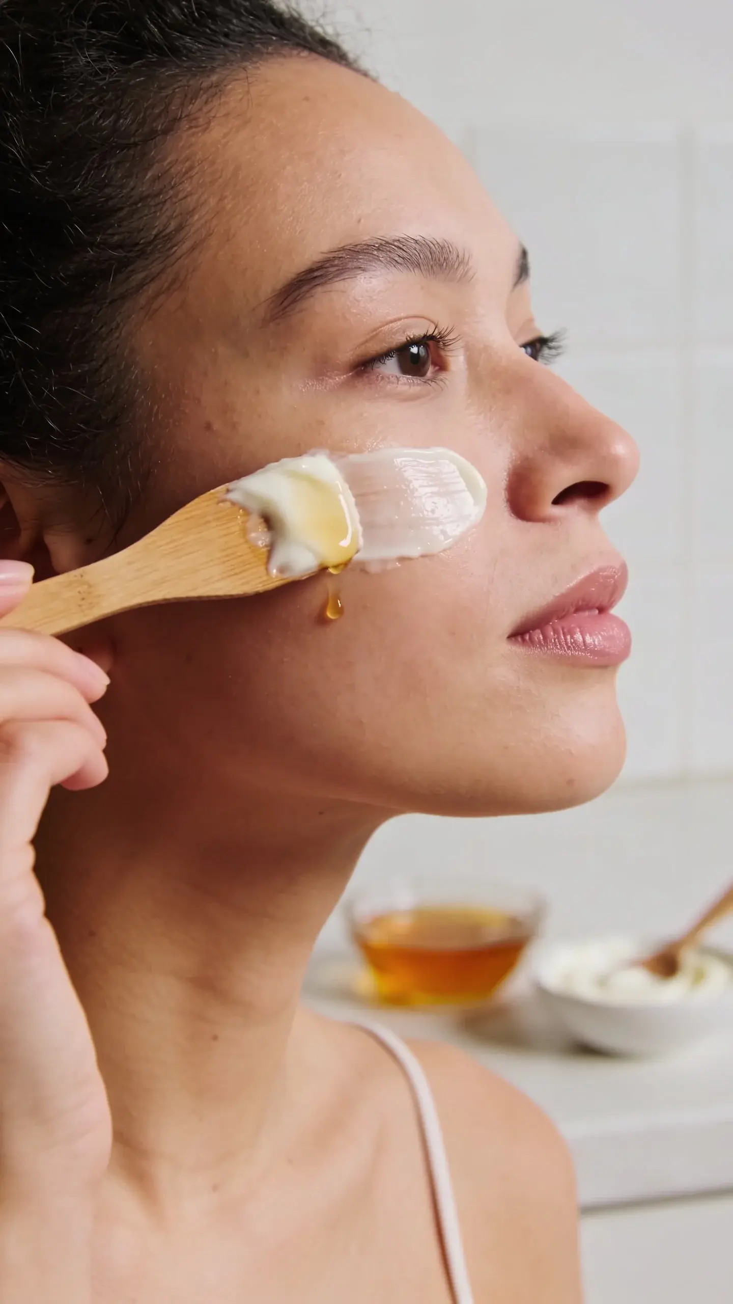 Close-up of a woman with smooth, glowing skin applying a creamy honey-yogurt face mask with a wooden spatula in a bright, minimal kitchen. Soft natural light, neutral tones, a small bowl of honey and a spoon of yogurt blurred in the background.