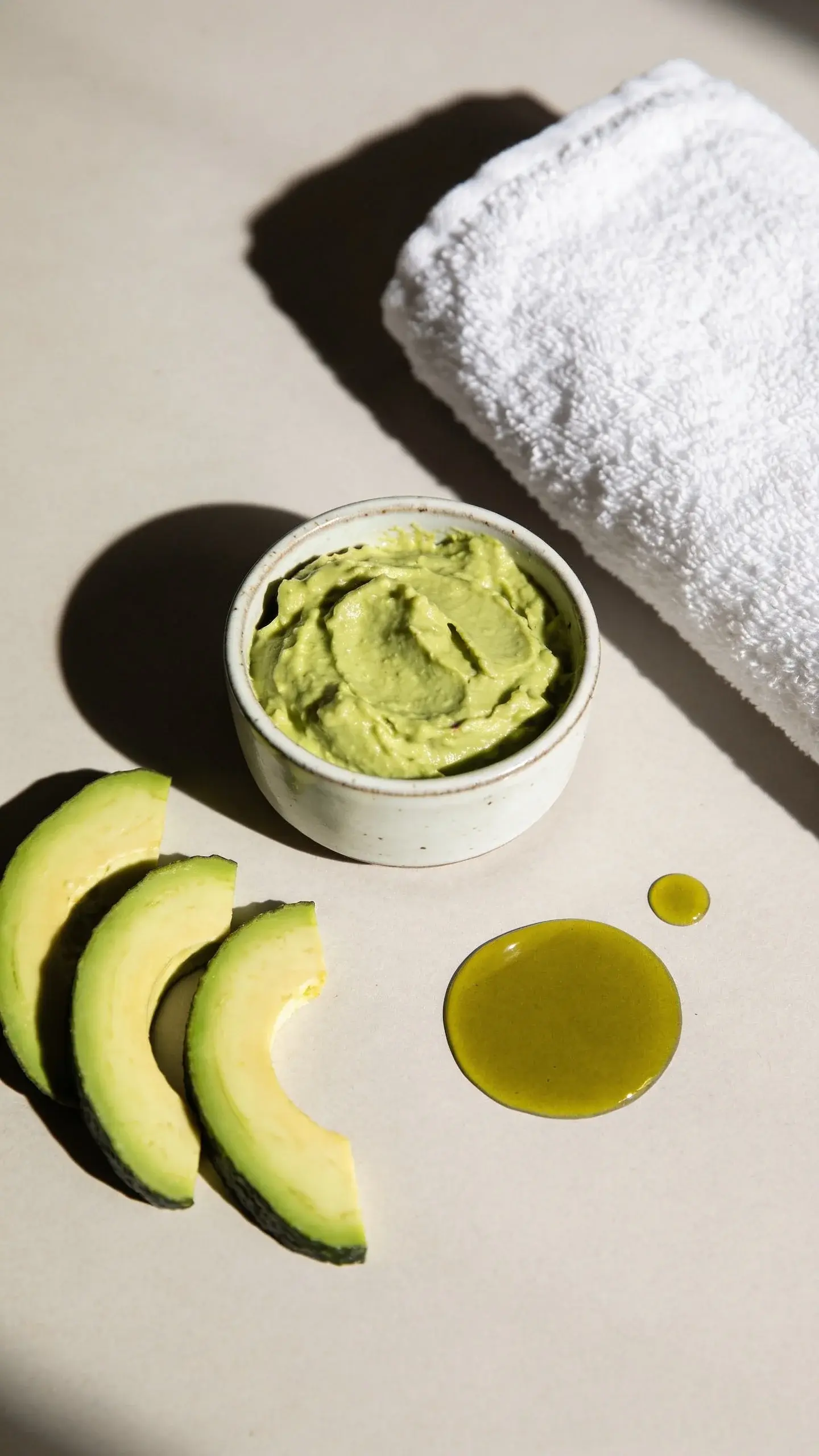 Overhead shot of a simple spa setup: a small ceramic bowl of mashed avocado mask beside slices of avocado, a drizzle of olive oil, and a soft white towel. Clean, airy aesthetic with natural light and gentle shadows, no text or labels.