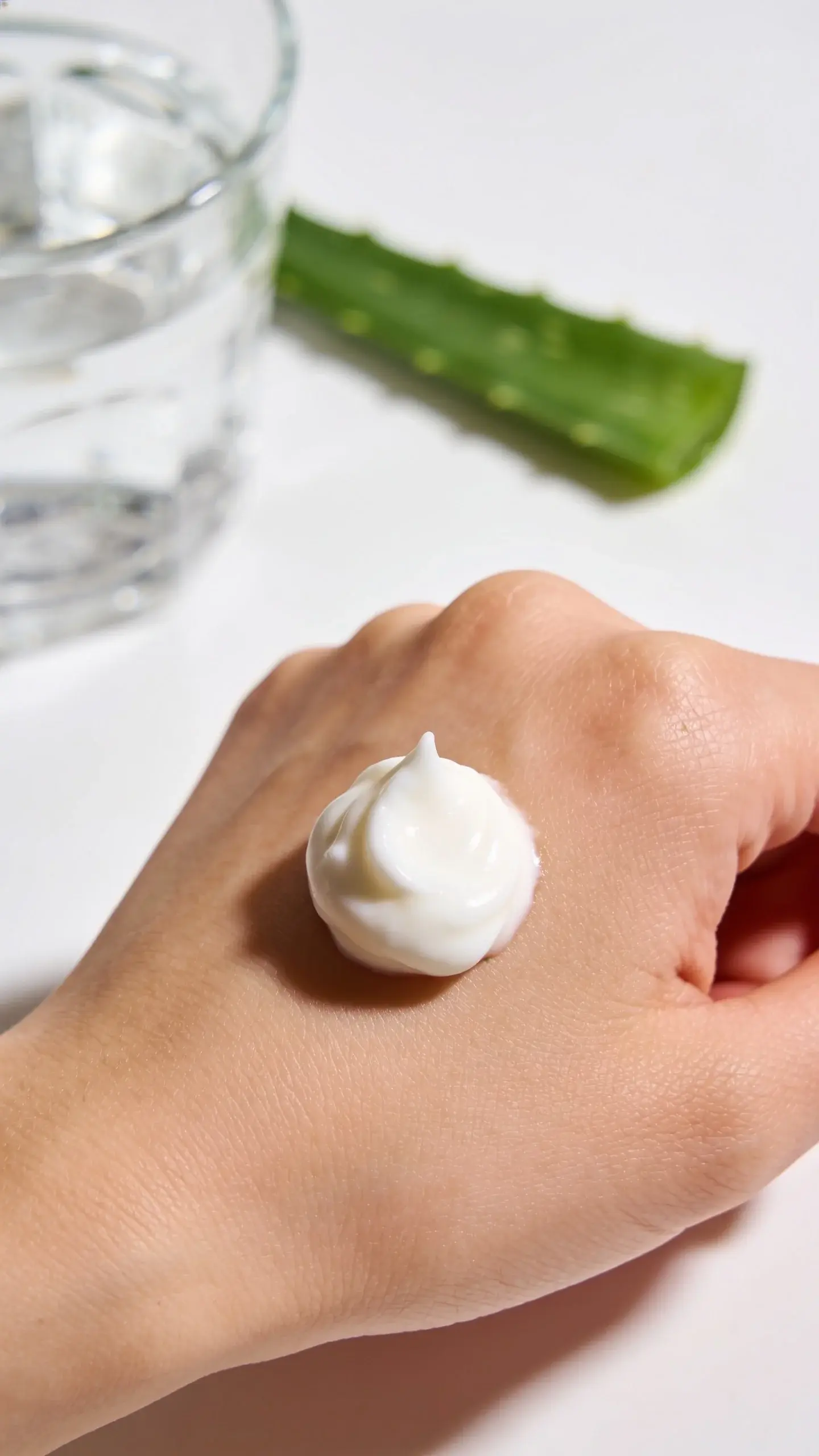 Overhead shot of a small scoop of thick, velvety face cream on the back of a clean hand, with a soft-focus glass of water and a sprig of aloe in the background. Bright, diffused light, clean white surface, emphasis on creamy texture and hydration vibes.