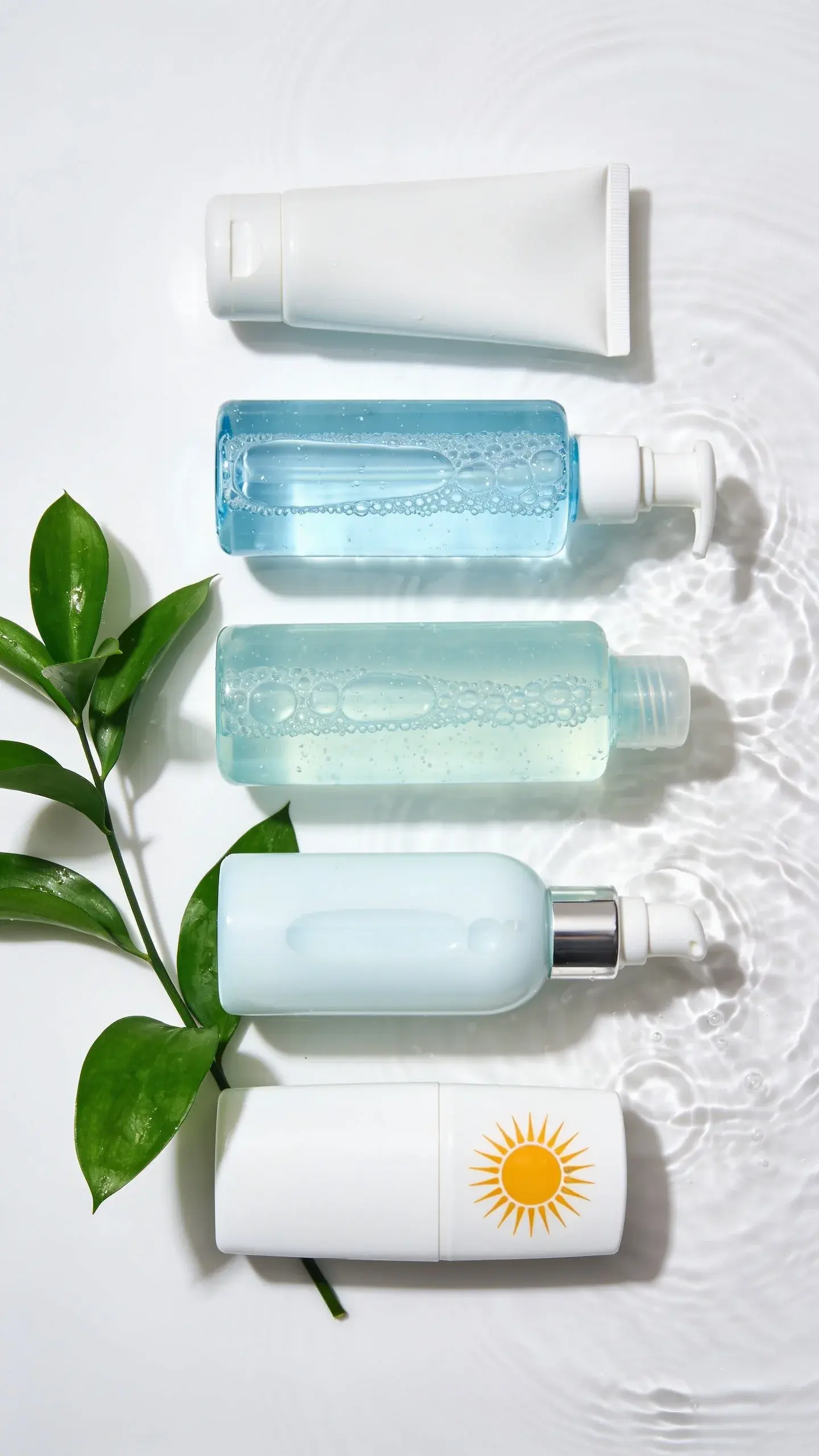 Flat lay of a beginner-friendly Korean skincare routine on a clean white surface: a gentle cleanser, watery toner, hydrating essence, light gel moisturizer, and sunscreen in sleek, unbranded containers. Add a sprig of green leaves and a ripple of water for a fresh, hydrating vibe, bright natural lighting.