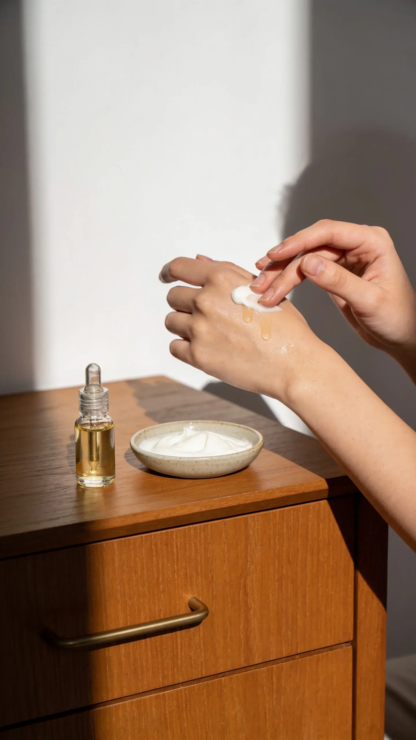 Close-up of hands applying a creamy moisturizer and a few drops of hydrating serum on smooth skin; soft daylight, glass dropper, and a simple ceramic dish on a wooden vanity. Warm, minimal aesthetic emphasizing hydration and barrier repair with dewy, healthy-looking skin.