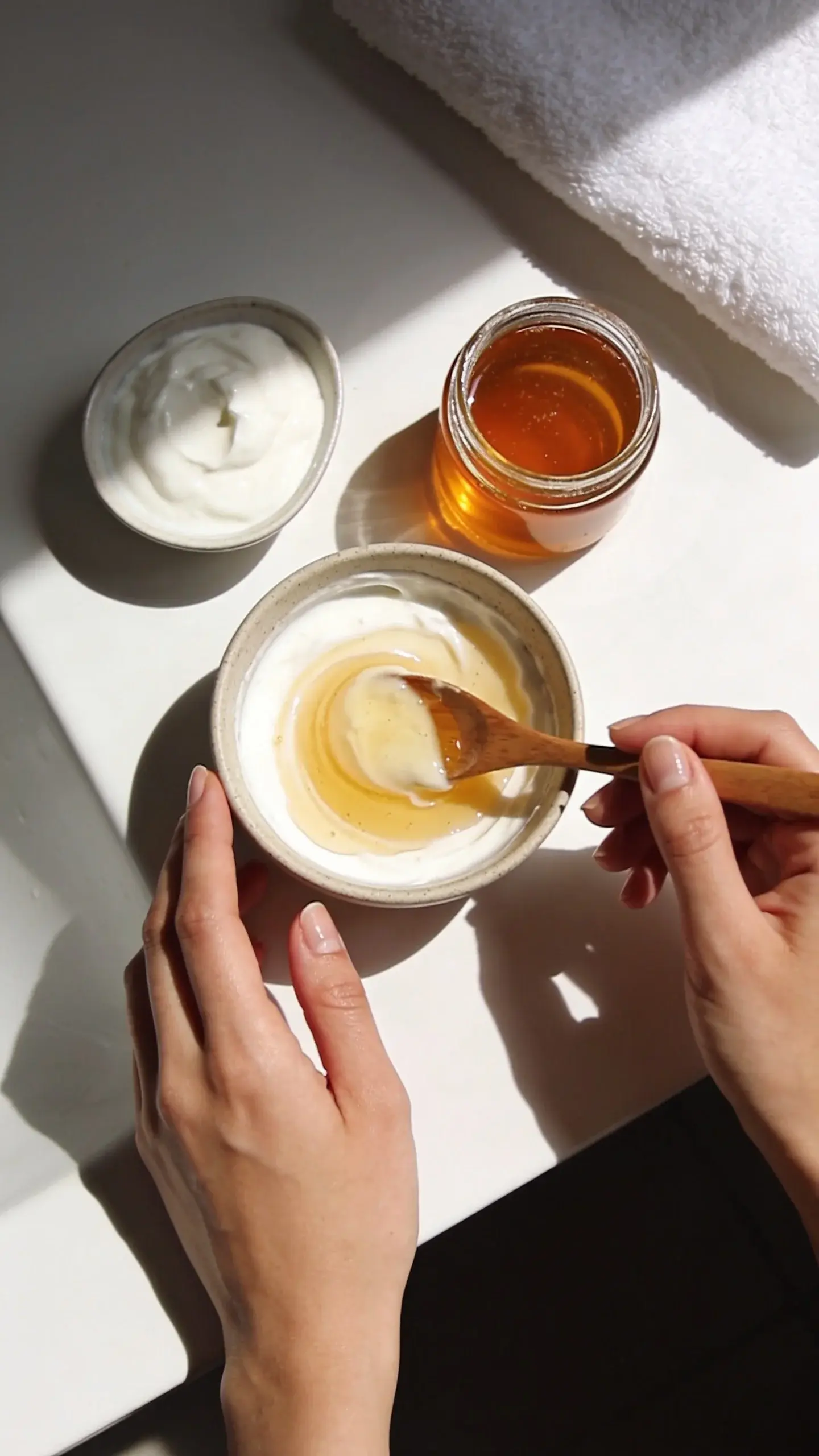 Overhead shot of a small ceramic bowl on a clean bathroom counter with a creamy honey and yogurt face mask mixture being stirred with a wooden spoon, beside a jar of honey, a small dish of plain yogurt, and a soft white towel; bright natural light, minimal, calm spa vibe, smooth healthy skin hands visible, no labels or text.