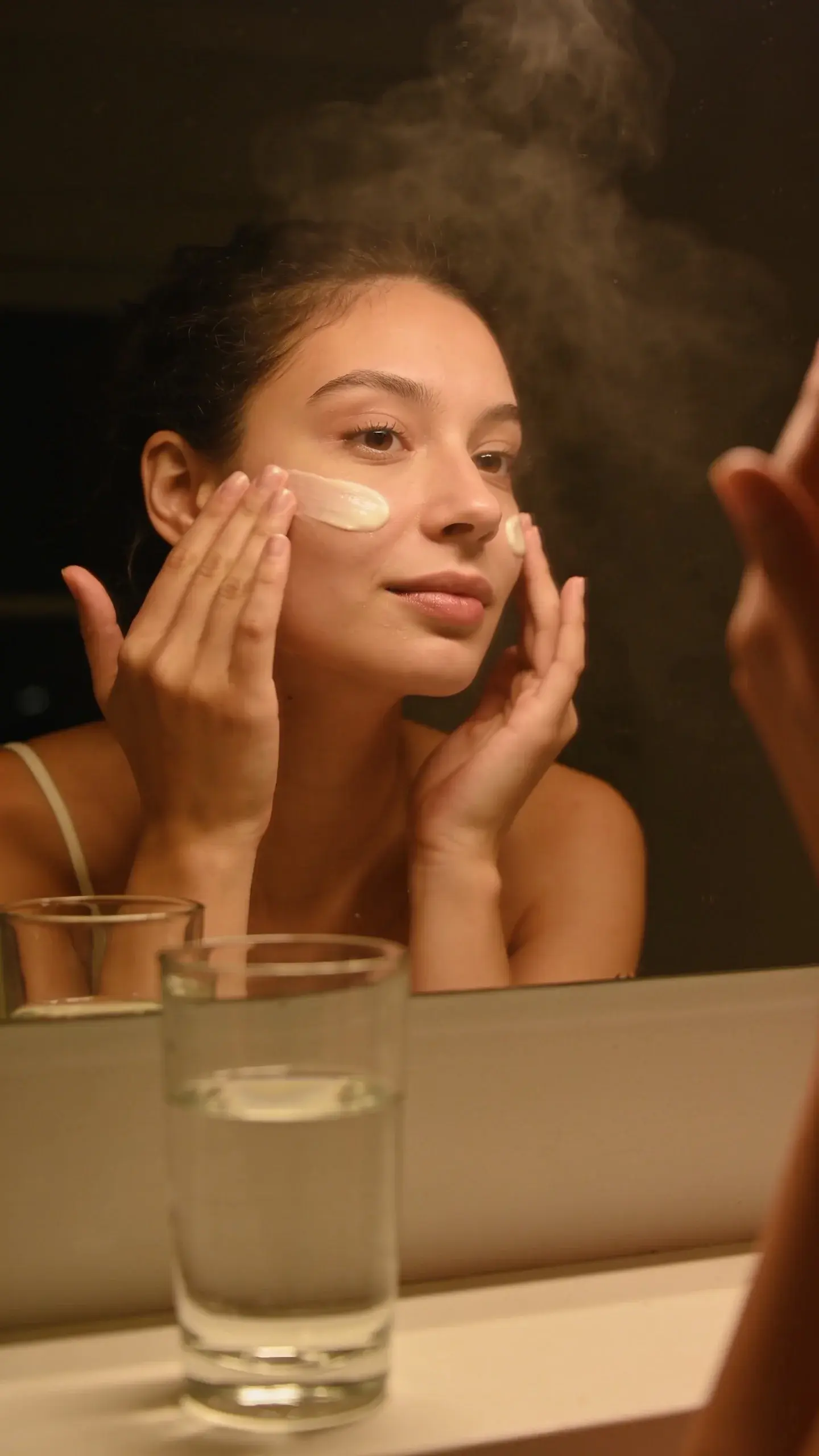 Cozy nighttime bathroom scene: a person applying a hydrating cream to their cheeks after cleansing, skin looking plump and smooth. Soft steam on the mirror, warm ambient lighting, simple neutral colors and a clean counter with a glass of water.