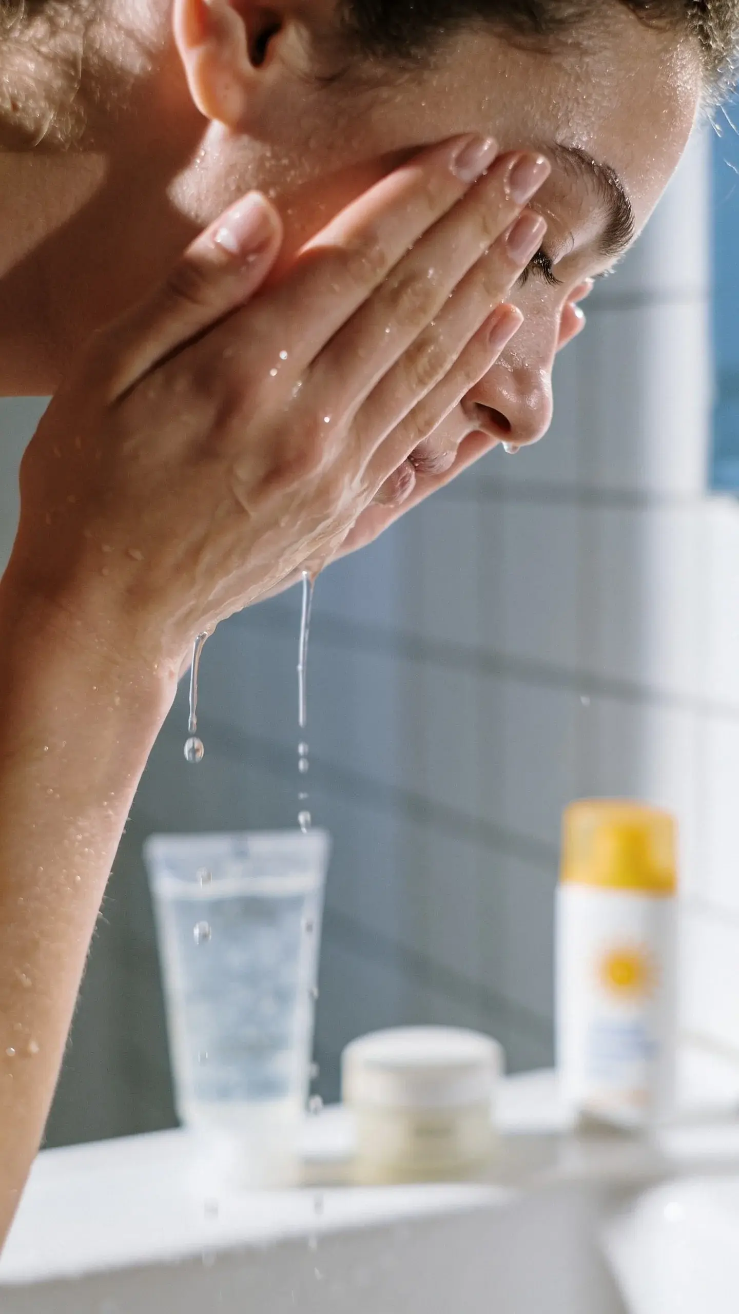 Soft morning bathroom scene with diffused natural light: a person with smooth, dewy skin gently patting water on their face, with a clear gel cleanser, a lightweight moisturizer, and a sunscreen bottle subtly in the background on a clean counter. Fresh, airy color palette with hints of white and pale blue, minimal clutter.