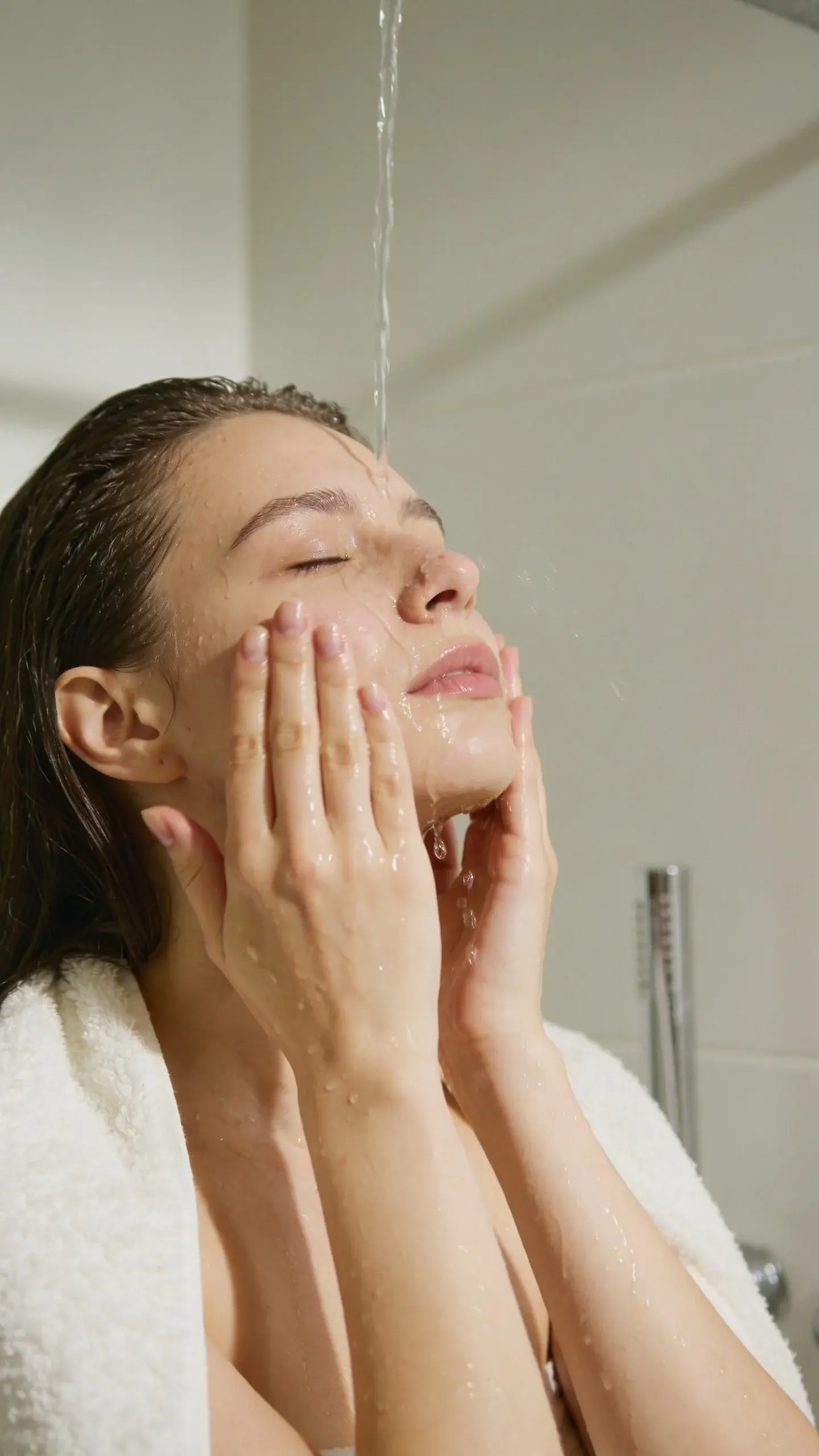 Close-up of a woman with smooth, dewy skin gently patting her face with water in a softly lit bathroom, a white fluffy towel draped over her shoulders, minimal and calming aesthetic with neutral tones and soft morning light.