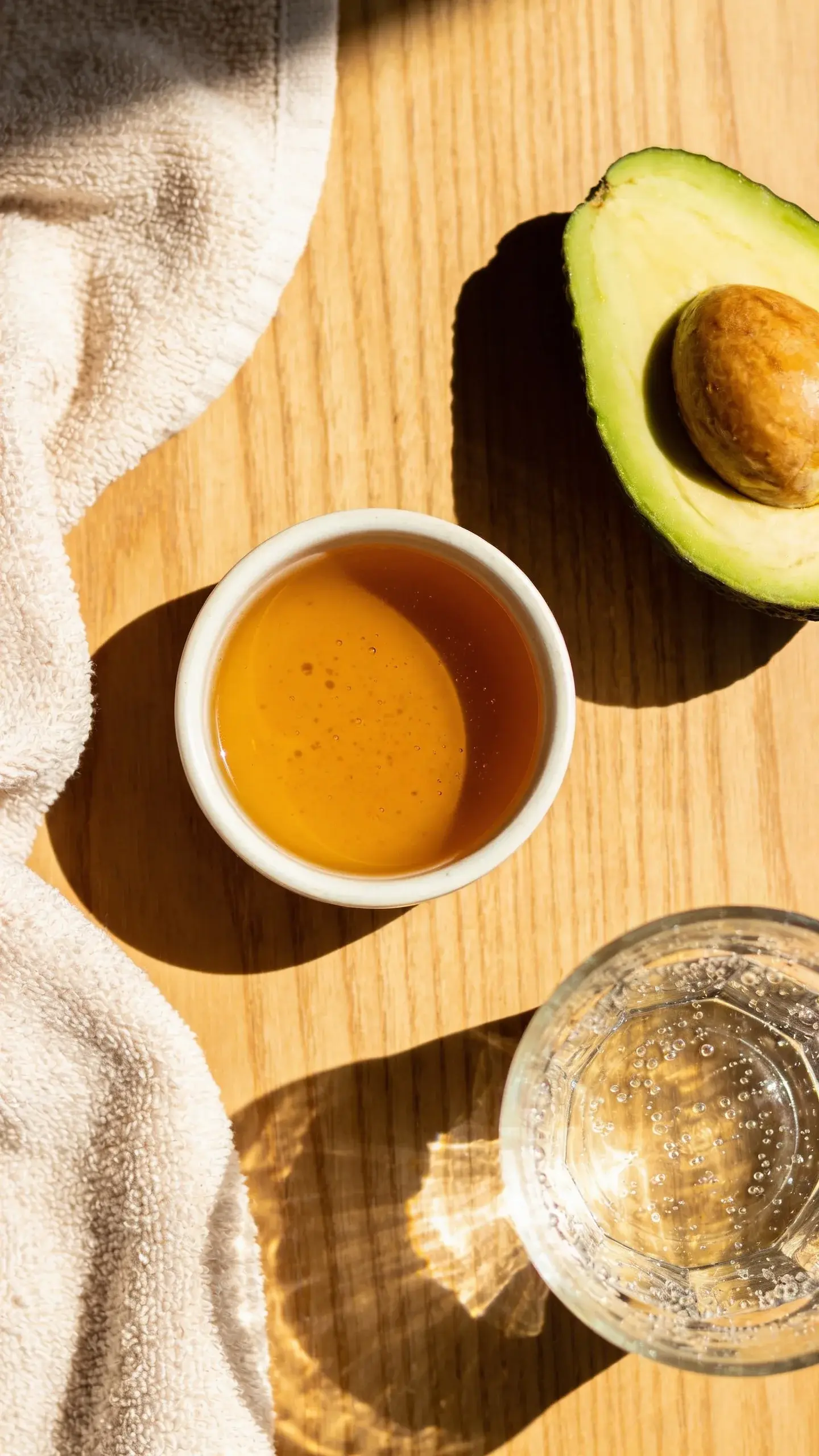 Overhead shot of a simple, natural skincare scene: a small bowl of honey, a sliced avocado, a glass of water with condensation, and a soft cotton towel on a clean wooden surface, warm natural light and a serene, spa-like vibe.