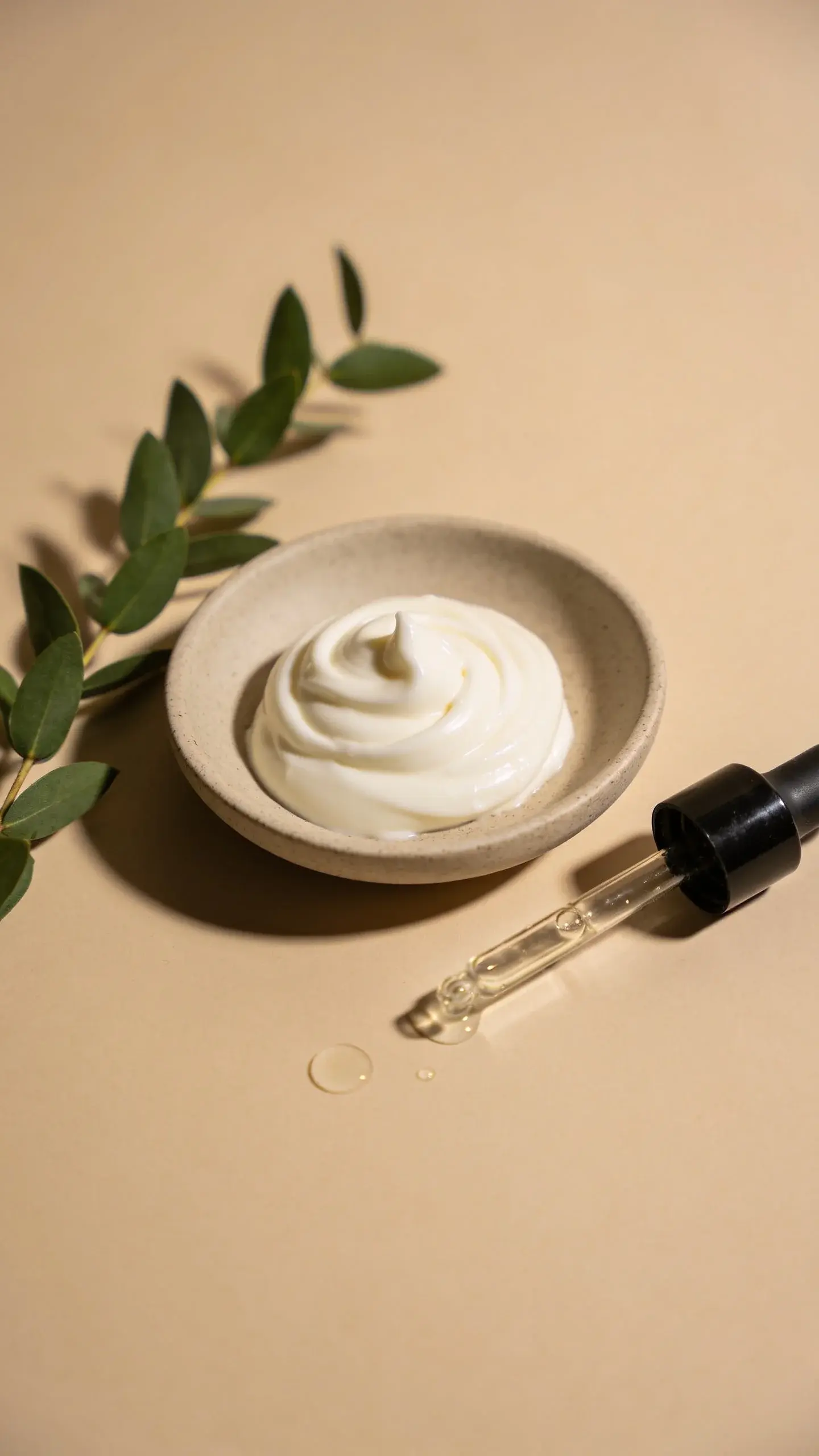 A simple flat lay of a small scoop of thick lotion swirled on a neutral ceramic dish beside a sprig of green leaves and a glass dropper with clear serum droplets; soft diffuse lighting, warm beige background, cozy spa vibe, no branding or text.