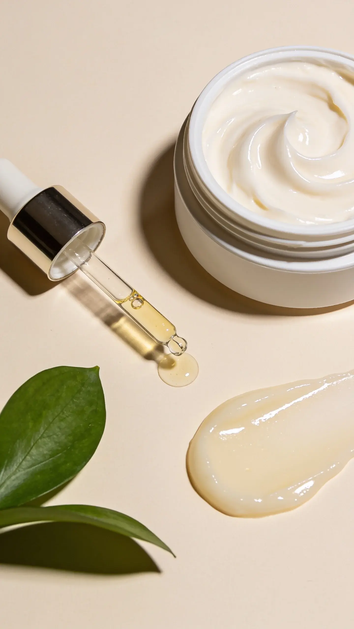 A clean, minimal flatlay of skincare essentials for dry skin on a soft beige background: an open jar of rich cream with a glossy swirl, a dropper bottle of serum with a droplet, and a dab of balm glistening. Soft natural light, subtle shadows, fresh green leaf accent, no labels or text.
