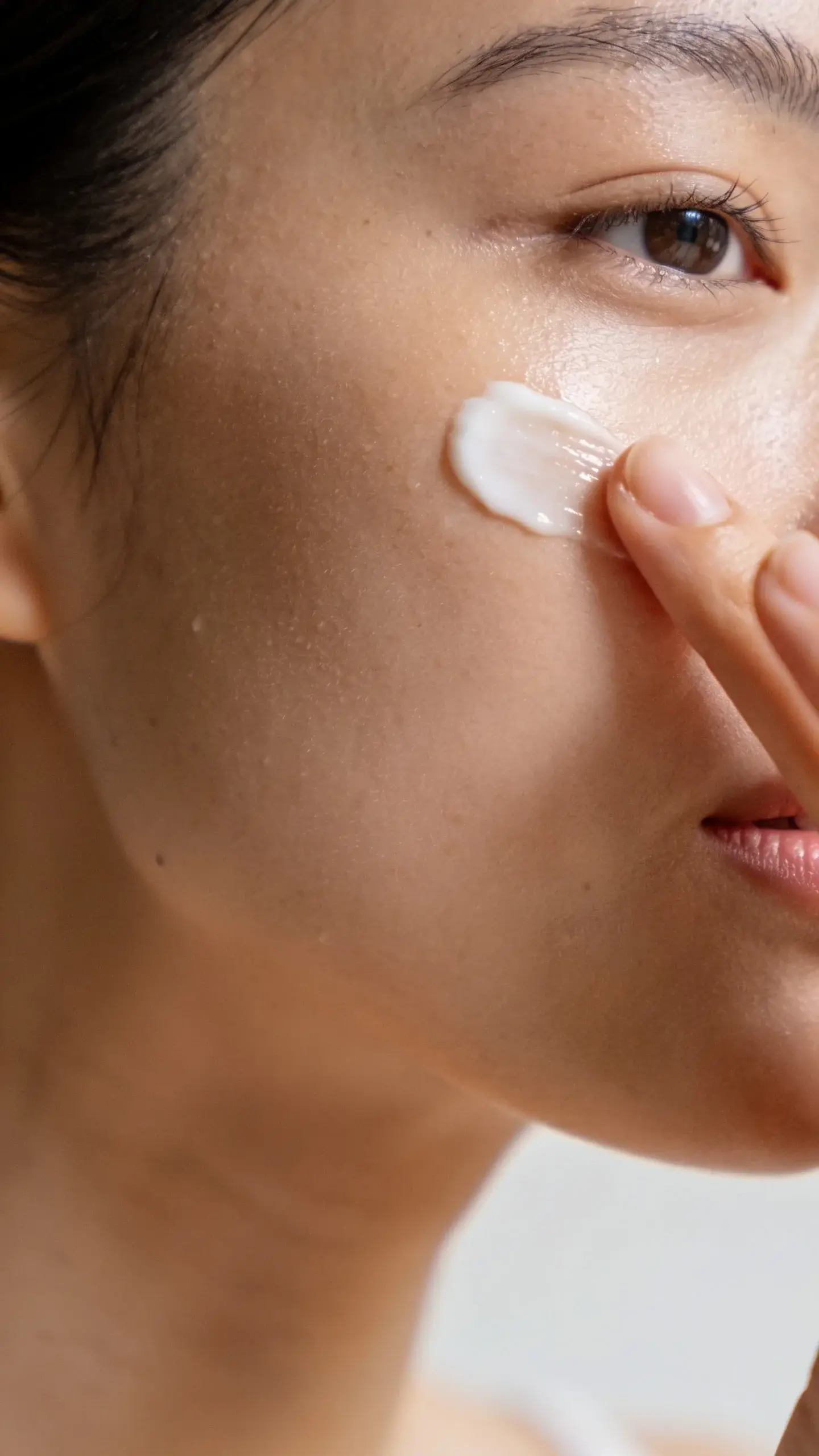 Close-up of smooth, dewy cheek and jawline with a fingertip gently applying a creamy moisturizer, catching soft morning light. Neutral background, luminous skin glow, shallow depth of field, no visible branding or text.