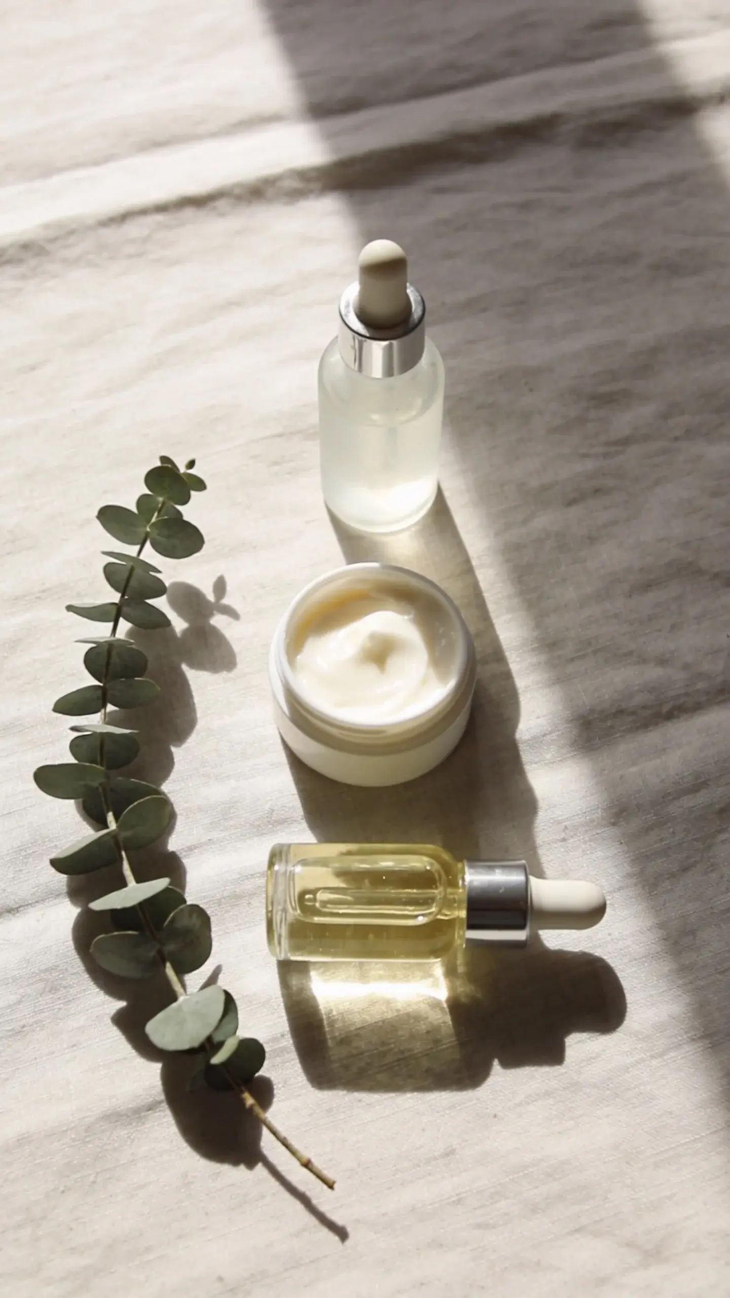 Flat lay on a light linen surface: a small dropper of translucent serum, a creamy balm, and a lightweight oil arranged in a simple sequence to suggest humectant → emollient → occlusive; soft morning light, neutral tones, and a sprig of eucalyptus for a calm, hydrated vibe.
