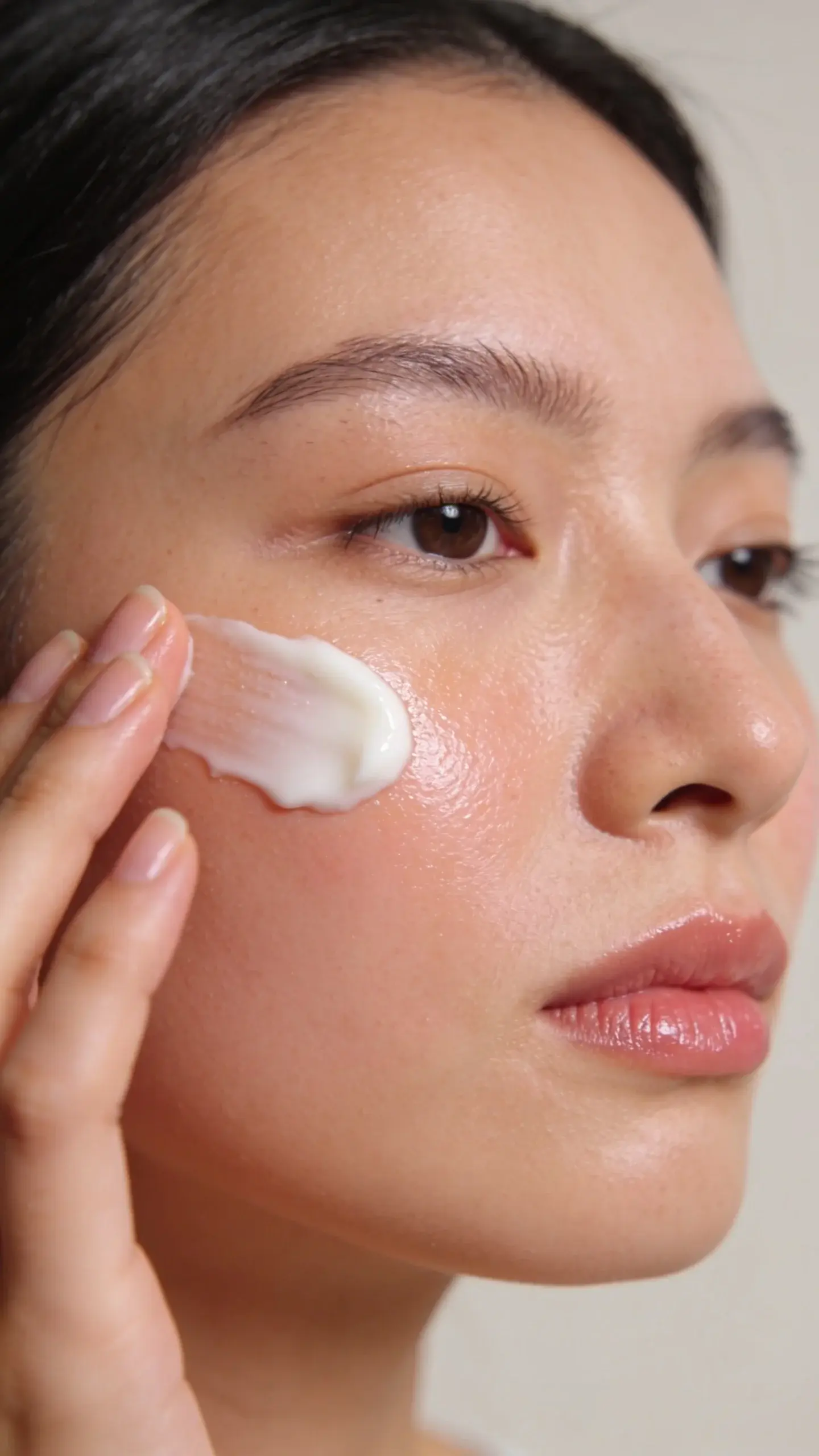 Close-up of a woman with smooth, dewy skin applying a generous layer of creamy sunscreen, soft natural daylight by a window, subtle glow on cheeks, minimal makeup, neutral background, focus on hydrated texture with no white cast.