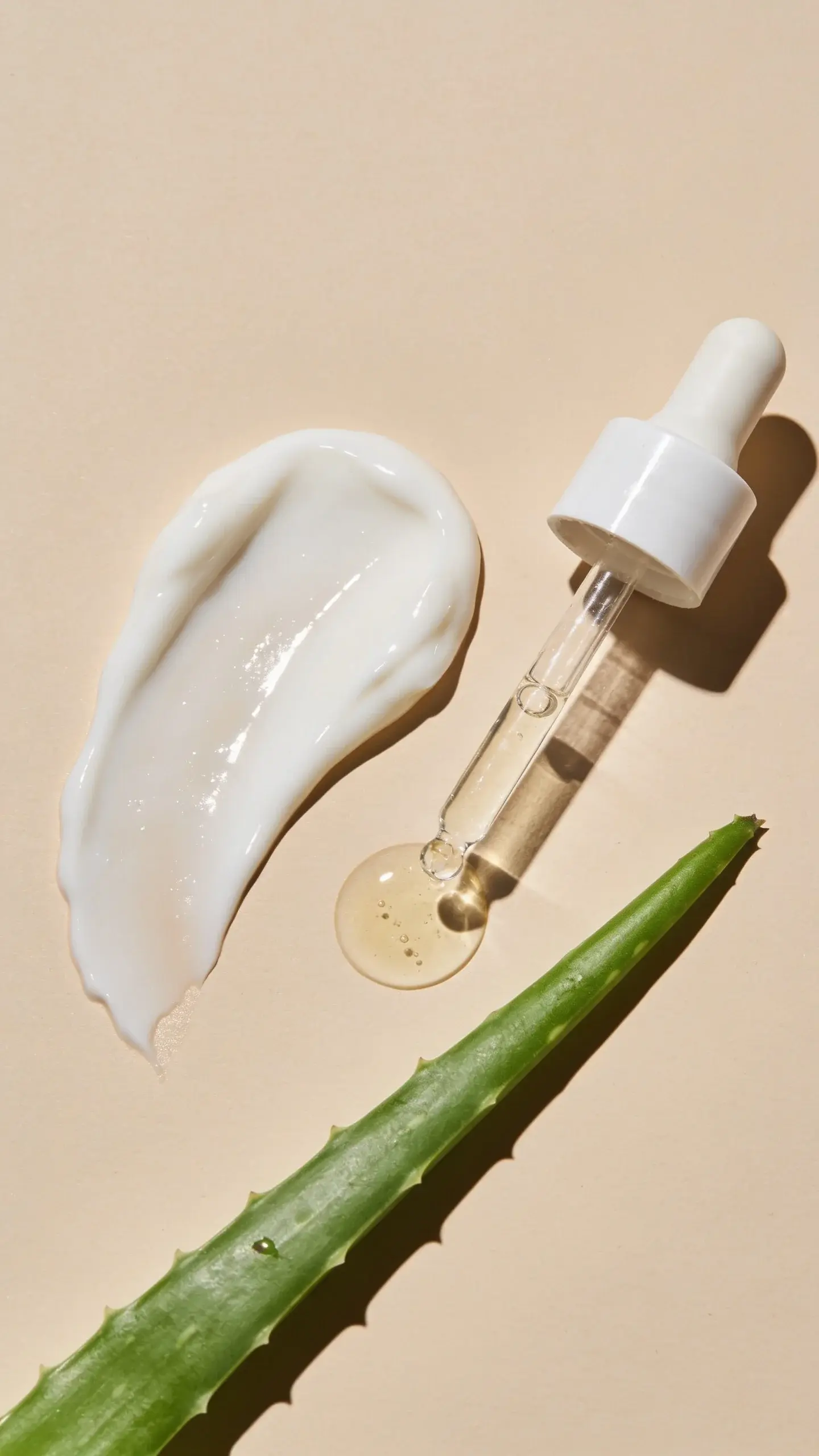 Flat lay of a glossy, milky sunscreen dollop beside a clear dropper of squalane and a sprig of aloe on a clean beige surface, soft shadows, sunlit highlight, overall fresh, hydrated vibe with a dewy sheen.