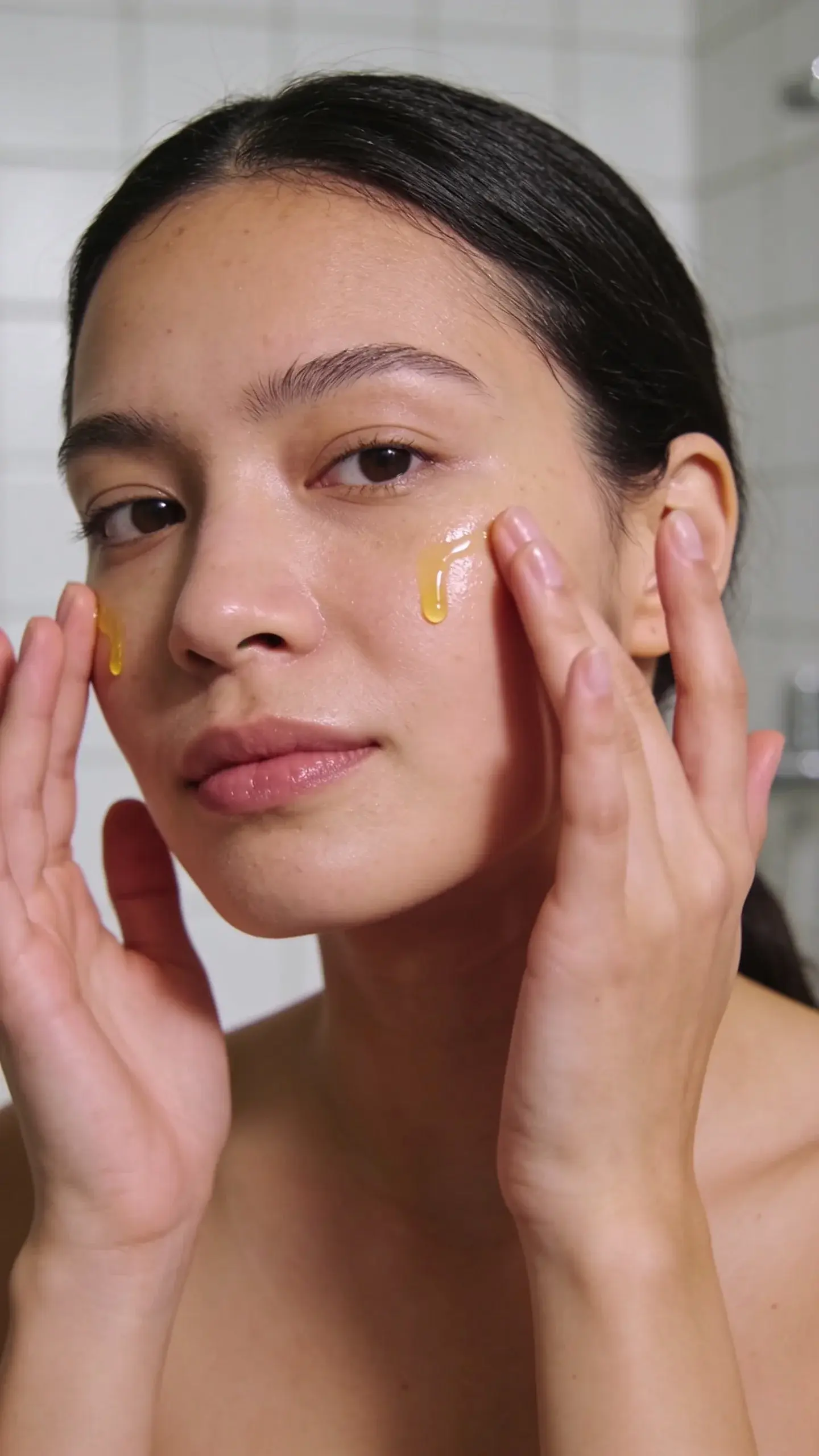 Close-up of a woman with smooth, dewy skin in soft morning light, gently pressing a few drops of facial oil into her cheeks with clean hands. Minimal bathroom background, neutral tones, subtle glow, no visible product labels, calm and fresh mood.