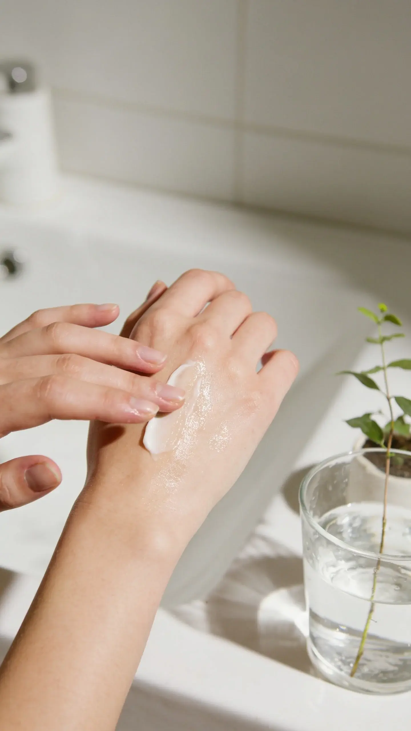 Close-up of a person gently applying a creamy moisturizer to smooth, healthy-looking skin in soft natural bathroom light, with a simple scene showing a glass of water and a small plant nearby to suggest hydration and calm. Neutral tones, minimal background, dewy finish on skin, no visible product labels.